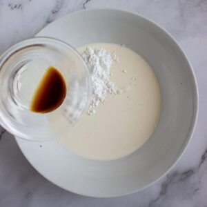 A white bowl contains cream, powdered sugar, and a splash of vanilla extract being poured in from a glass on a marble countertop.