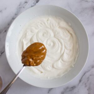 A spoonful of peanut butter is held above a bowl of whipped cream mixture on a marble surface.