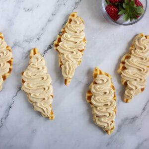 Five waffle pieces topped with swirled cream are arranged on a marble surface; a bowl of strawberries is partially visible in the corner.