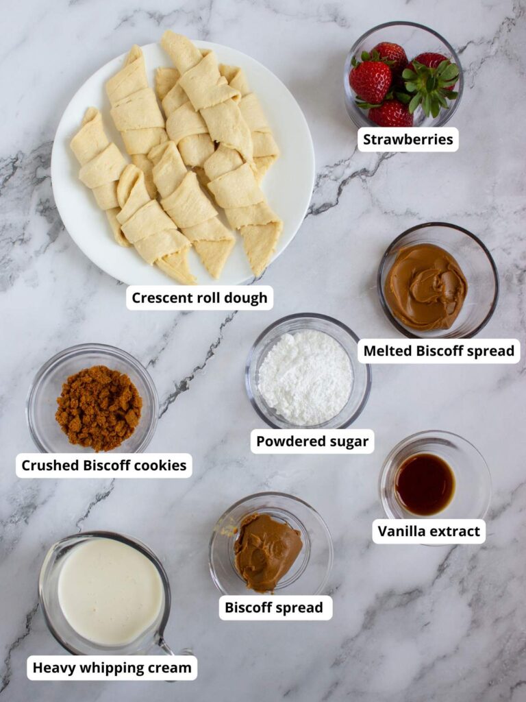 Ingredients for a dessert are arranged on a marble surface, including crescent roll dough, strawberries, Biscoff spread, melted Biscoff, crushed cookies, powdered sugar, vanilla, and cream.
