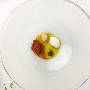 Clear glass bowl with small piles of paprika, onion powder, salt, black pepper, and olive oil arranged inside.