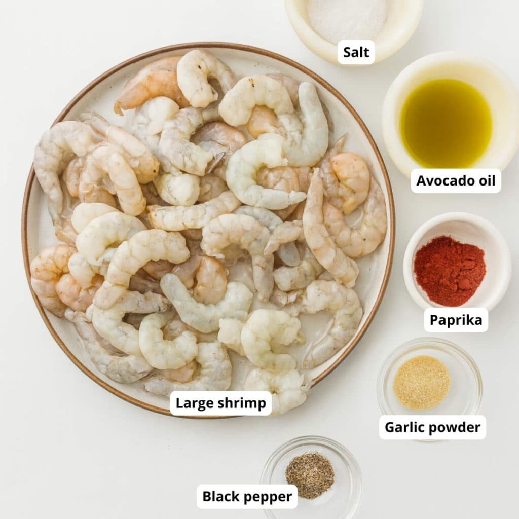 A plate of raw large shrimp is surrounded by bowls of salt, avocado oil, paprika, garlic powder, and black pepper.