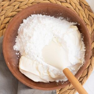 Brown bowl with powdered sugar and cream cheese being mixed with a spatula, placed on a woven placemat.