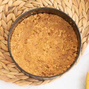 Round pan filled with a pressed, crumbly biscuit base sits on a woven placemat, viewed from above.