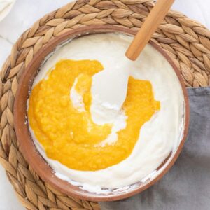 A mixing bowl with white cream and yellow purée being stirred with a spatula, placed on a woven placemat with a gray napkin nearby.
