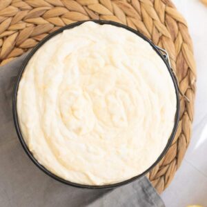 A round pan filled with creamy, swirled batter sits on a woven placemat with a corner of a gray cloth visible.