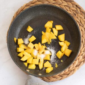 Chopped mango pieces and sprinkled sugar in a black frying pan on a woven placemat.