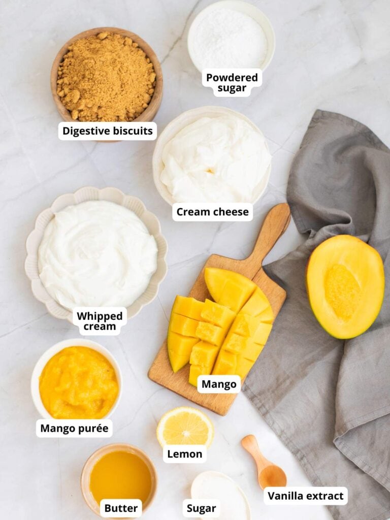 Various labeled ingredients for a mango cheesecake are arranged on a white surface.