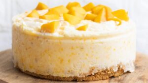 A round mango cheesecake with a crumbly graham cracker crust is topped with chunks of fresh yellow mango, placed on a wooden board.