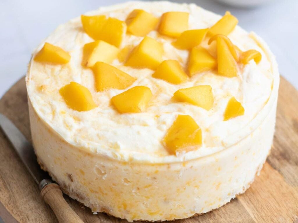 A round cheesecake topped with chunks of yellow mango sits on a wooden board next to a knife.