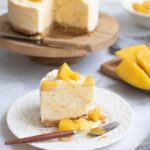 A slice of mango cheesecake on a white plate with a fork, with the whole cheesecake and sliced mangoes in the background.