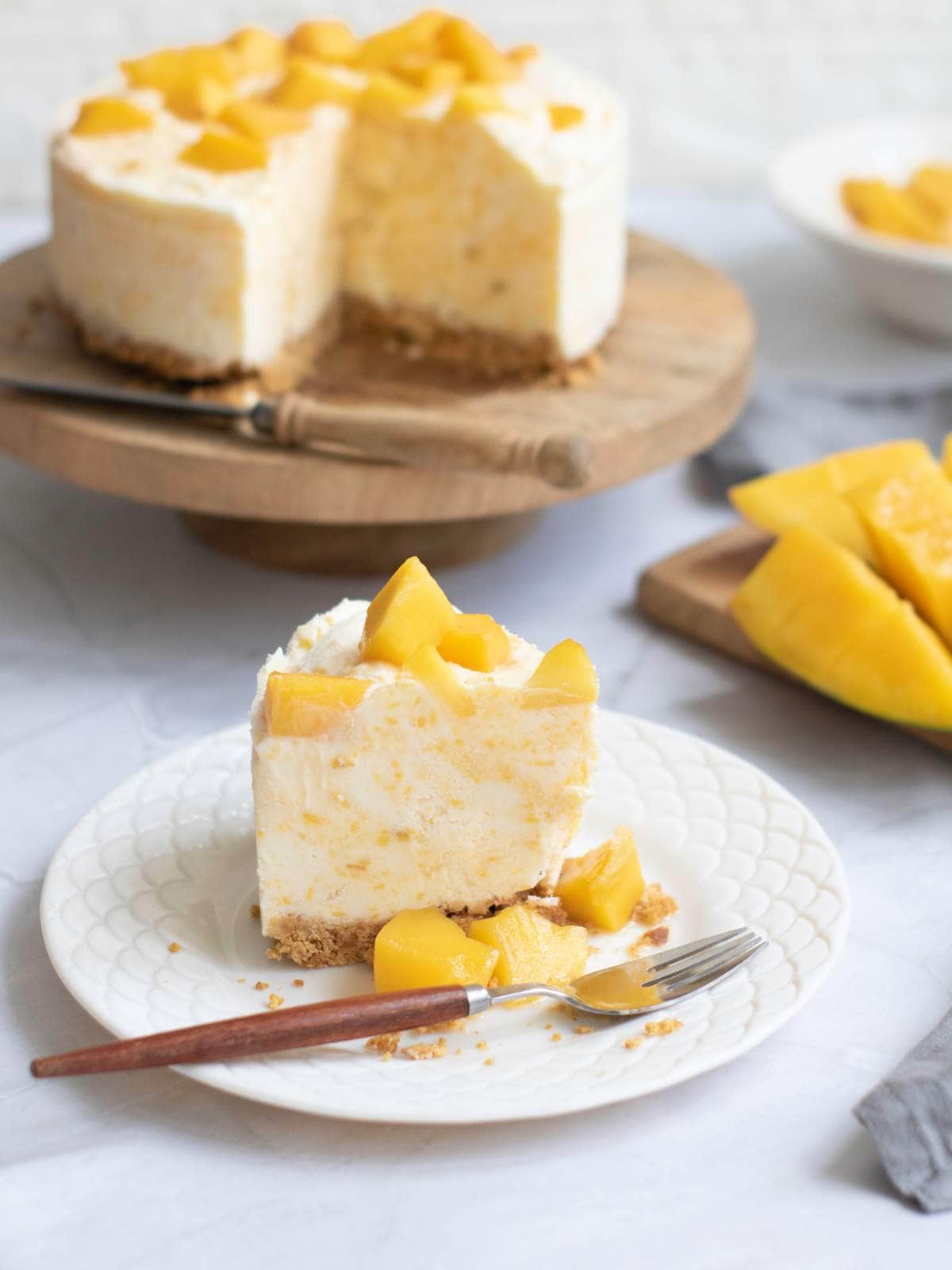 A slice of mango cheesecake on a white plate with a fork, with the whole cheesecake and sliced mangoes in the background.