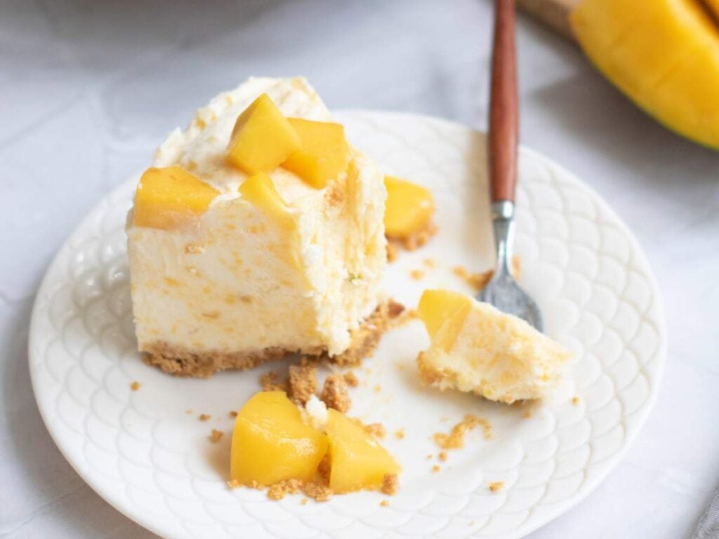 A slice of cheesecake with a graham cracker crust and mango pieces on a white plate, next to a fork and a few crumbs.