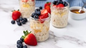 Three small jars of overnight oats topped with strawberries and blueberries, with additional fruit scattered nearby and a small bowl of honey in the background.