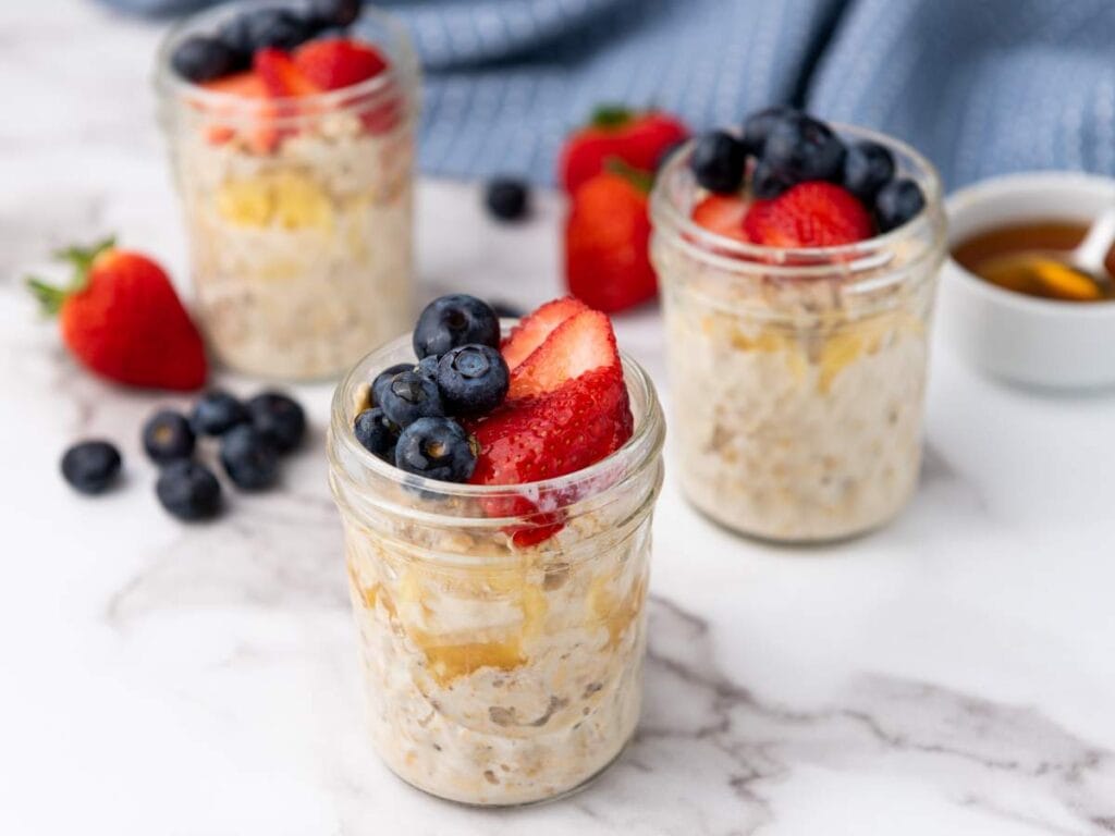 Three small jars of overnight oats topped with sliced strawberries and blueberries sit on a marble surface, with extra berries and a bowl of honey in the background.