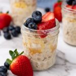 A small glass jar of overnight oats topped with fresh strawberries and blueberries sits on a marble surface, with more berries scattered nearby.