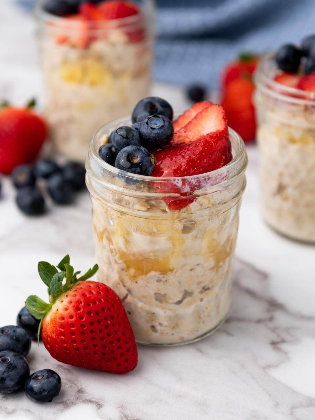 A small glass jar of overnight oats topped with fresh strawberries and blueberries sits on a marble surface, with more berries scattered nearby.