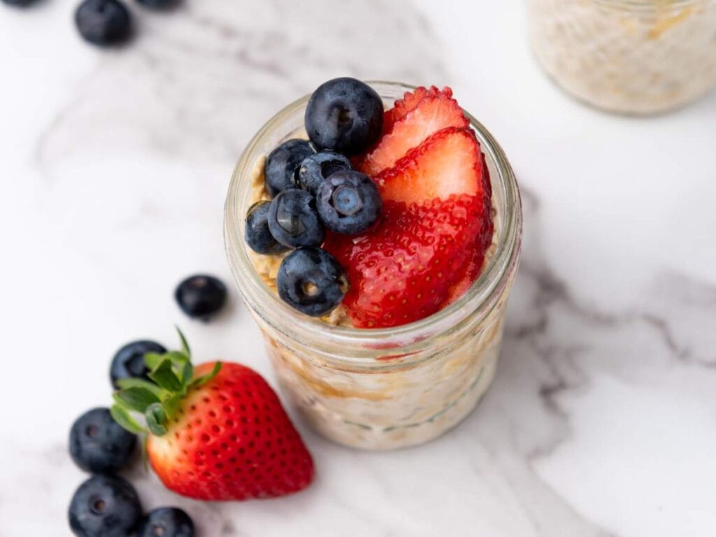 A glass jar of overnight oats topped with sliced strawberries and blueberries, with additional berries scattered on a marble surface nearby.