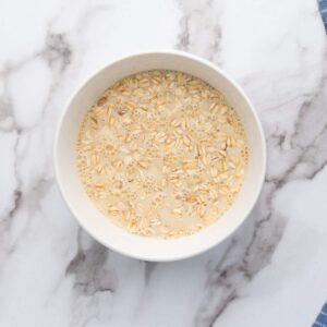 A white bowl filled with raw oats soaking in milk, placed on a white and gray marble surface.