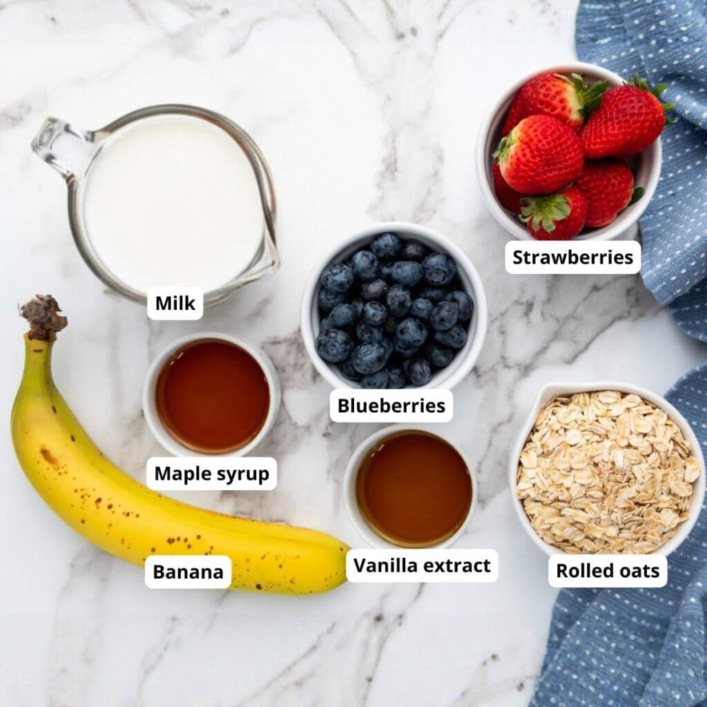 A banana, strawberries, blueberries, rolled oats, milk, maple syrup, and vanilla extract arranged on a marble surface.