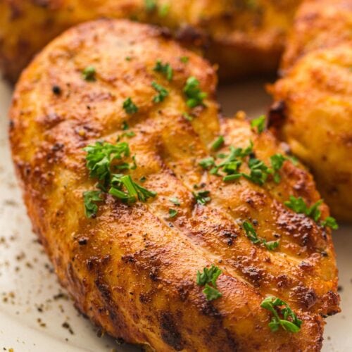 Grilled seasoned chicken breast garnished with chopped parsley sits on a white plate.