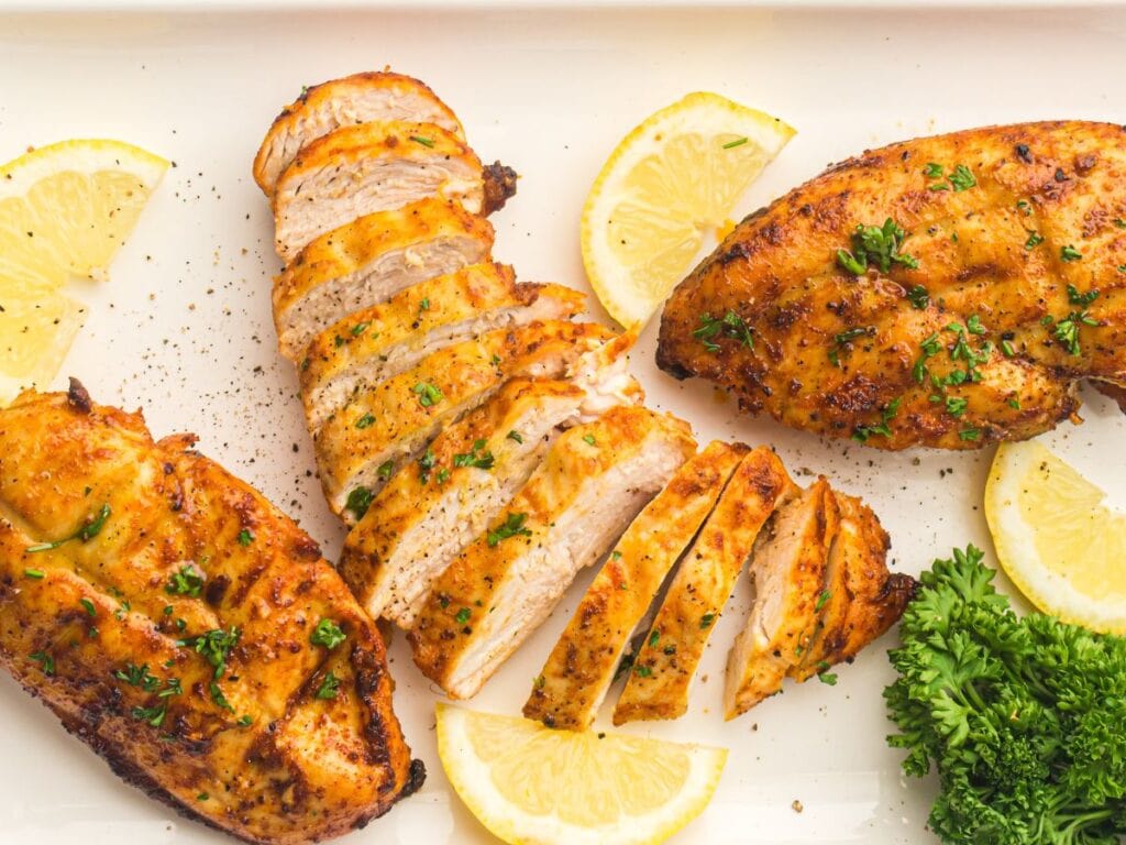 Grilled chicken breasts garnished with herbs, served with lemon wedges and parsley on a white plate.