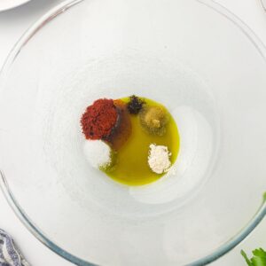 A glass bowl containing olive oil, paprika, salt, black pepper, garlic powder, and brown sugar on a white surface.
