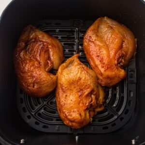Three uncooked, seasoned chicken breasts sit inside the basket of an air fryer, ready to be cooked.