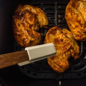 Three cooked pieces of seasoned chicken in an air fryer basket, with a pair of tongs holding one piece.