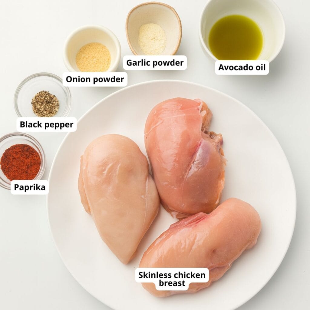 Three raw skinless chicken breasts on a plate, surrounded by small bowls of avocado oil, garlic powder, onion powder, black pepper, and paprika, each ingredient labeled.
