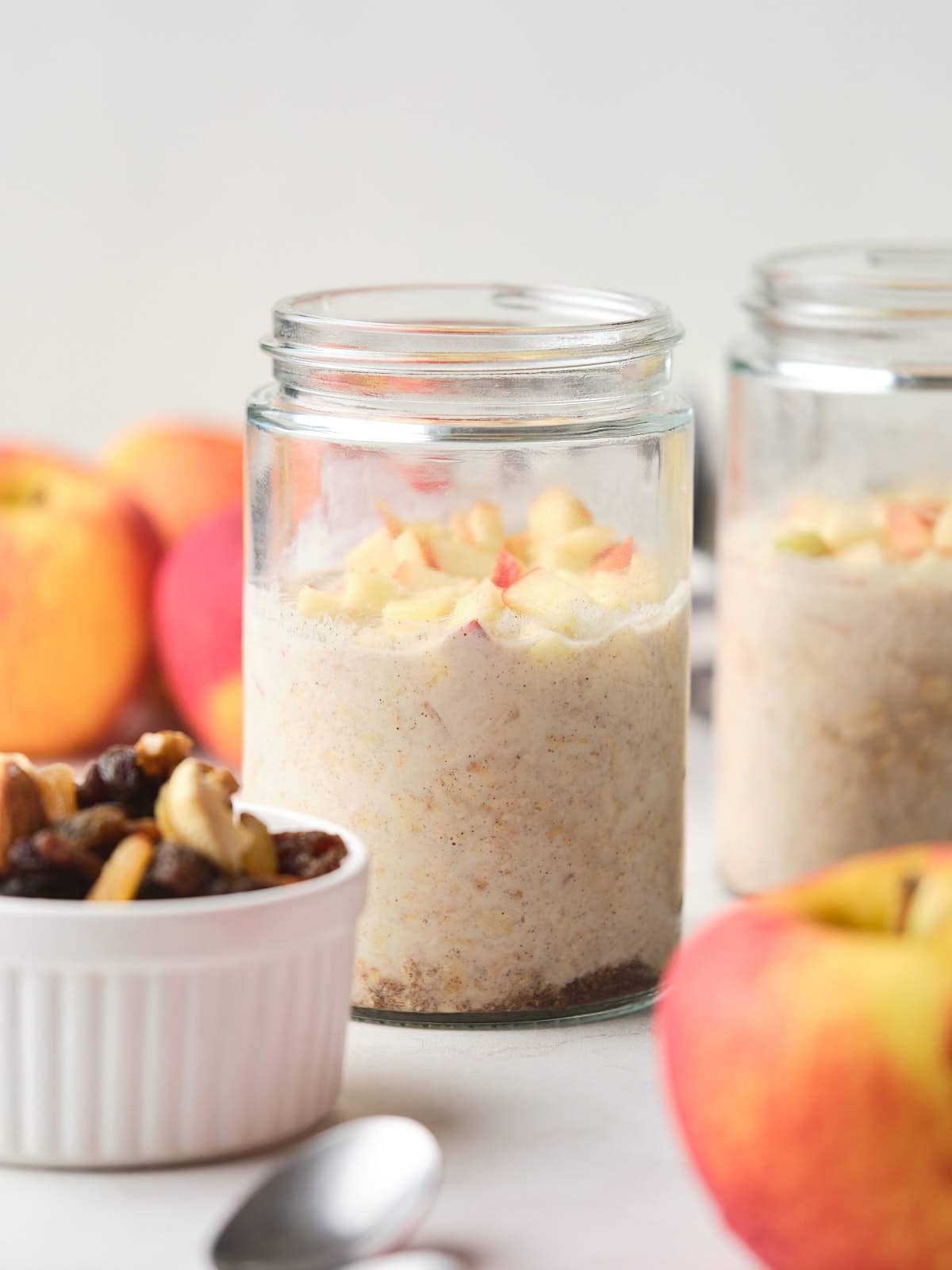 A glass jar of oatmeal topped with diced apples sits on a white surface, with apples and a small bowl of mixed nuts and raisins nearby.