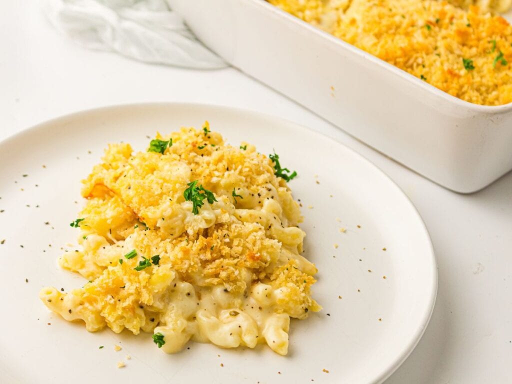 Baked macaroni and cheese topped with breadcrumbs and herbs is served on a white plate, with a baking dish of macaroni and cheese in the background.