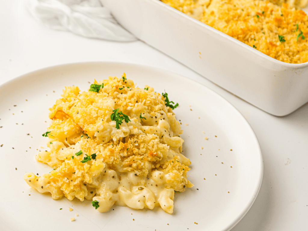 Baked macaroni and cheese topped with breadcrumbs and herbs is served on a white plate, with a baking dish of macaroni and cheese in the background.
