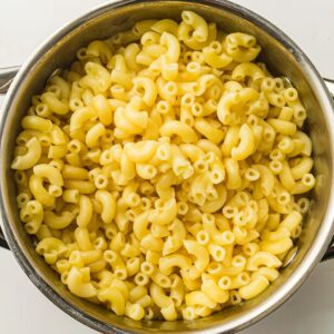 A stainless steel pot filled with cooked elbow macaroni pasta, viewed from above on a white surface.