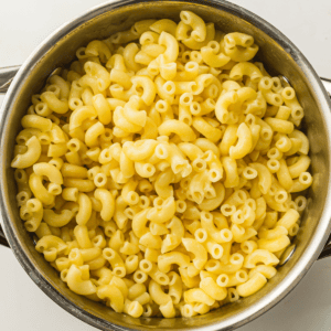 A stainless steel pot filled with cooked elbow macaroni pasta, viewed from above on a white surface.