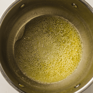 Close-up of melted butter with bubbles forming in a stainless steel saucepan, seen from above.