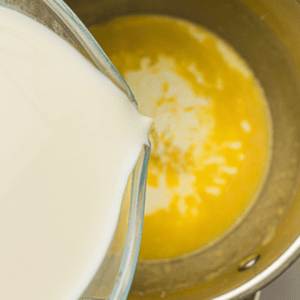 Milk is being poured from a glass container into a saucepan containing melted butter.