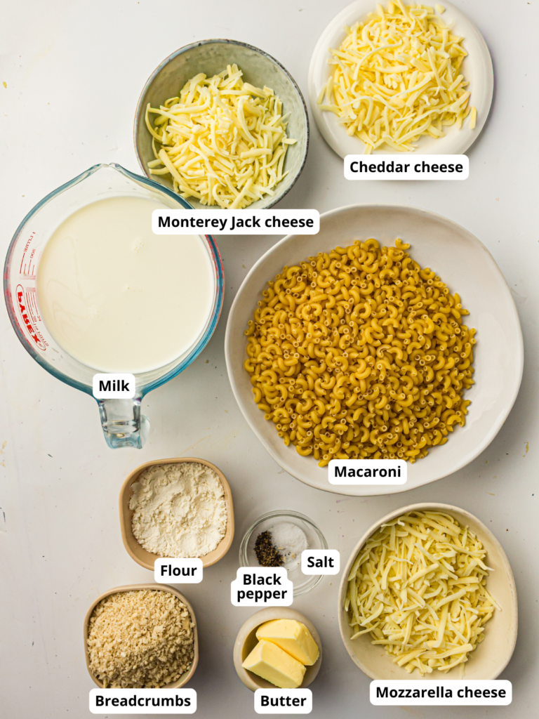 A top-down view of labeled ingredients for macaroni and cheese, including milk, macaroni, butter, flour, black pepper, salt, cheddar, mozzarella, Monterey Jack cheese, and breadcrumbs.