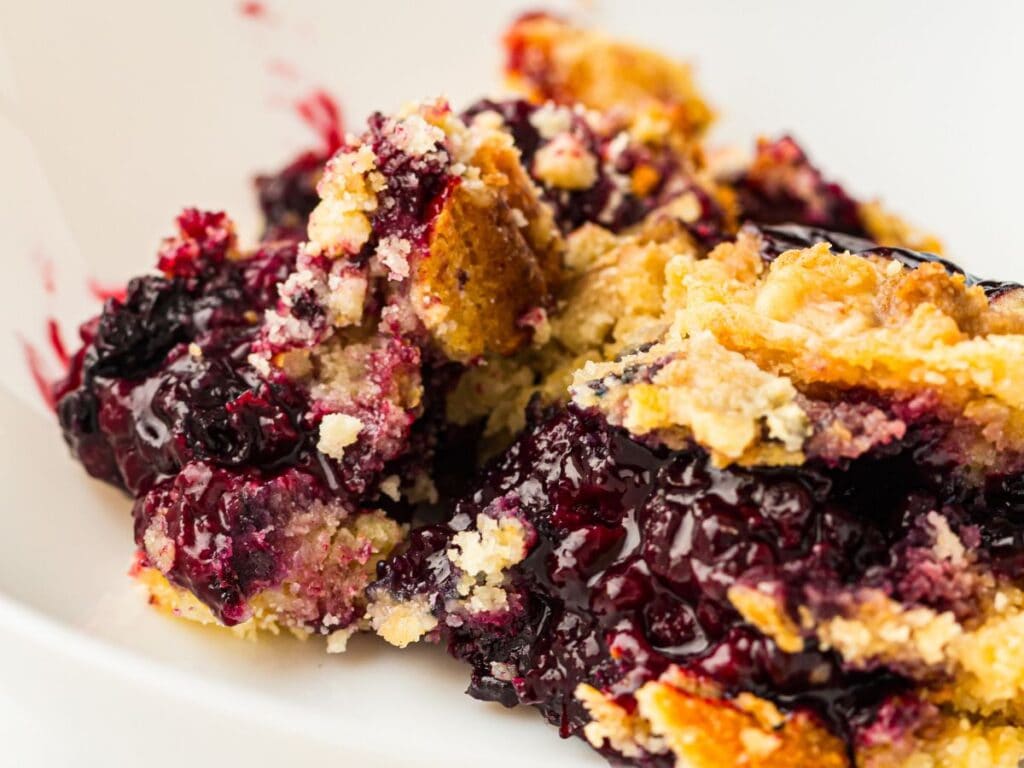 A close-up of a serving of blueberry pie with a golden, crumbly topping on a white plate.