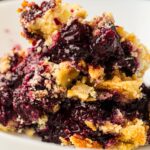A close-up of a serving of blueberry dump cake with a crumbly golden crust and juicy berry filling on a white plate.