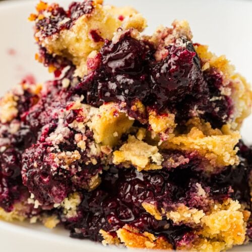 A close-up of a serving of blueberry dump cake with a crumbly golden crust and juicy berry filling on a white plate.