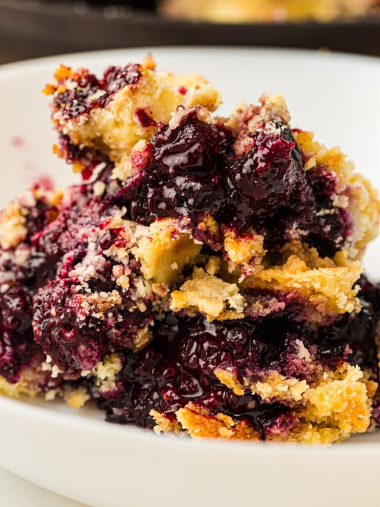 A close-up of a serving of blueberry dump cake with a crumbly golden crust and juicy berry filling on a white plate.