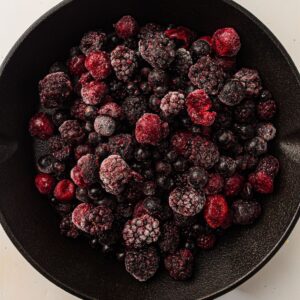 A cast iron pan filled with an assortment of frozen mixed berries, including blackberries, raspberries, and blueberries, covered in a light layer of frost.