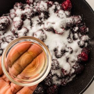 A hand holds a small bowl of liquid over a pan filled with blackberries coated in sugar.
