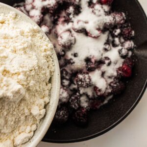 A bowl of flour next to a bowl of mixed berries topped with sugar, viewed from above.