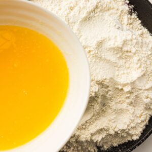 A bowl of melted butter next to a pile of flour on a dark surface, showing ingredients ready to be combined for baking.