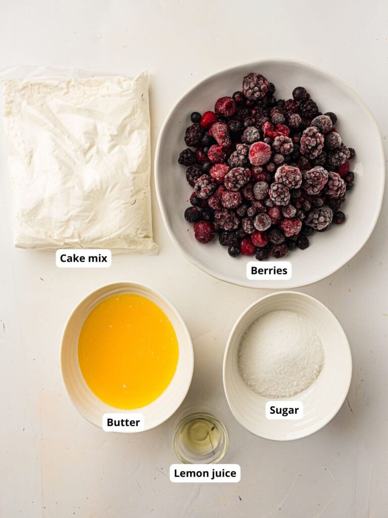 Five labeled bowls on a white surface containing cake mix, frozen berries, melted butter, sugar, and lemon juice.