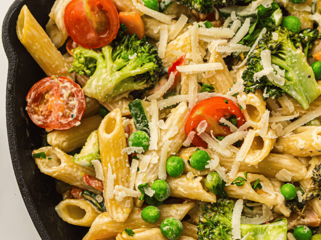 Close-up of penne pasta mixed with broccoli, cherry tomatoes, green peas, spinach, and topped with shredded cheese in a black pan.