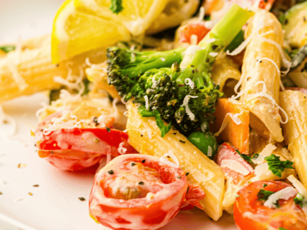 Close-up of penne pasta with broccoli, cherry tomatoes, shredded cheese, and black pepper, garnished with a lemon wedge.