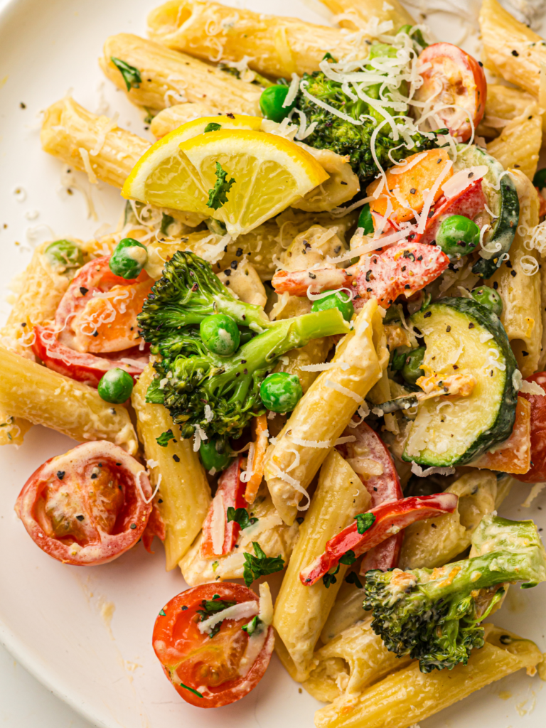 Penne pasta with broccoli, peas, zucchini, tomatoes, and red peppers, topped with grated cheese, lemon slices, and parsley on a white plate.